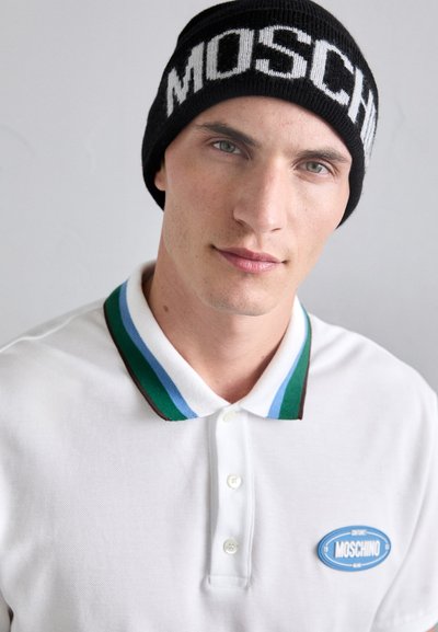 Mustrend mustekate, millel on valge "MOSCHINO". Valge polo särk sinise, rohelise ja pruuni triibuline kaelus ning sinine logotempel.