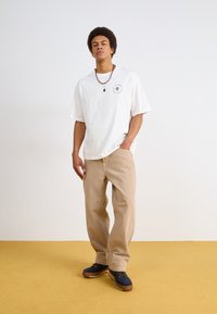 Hvit oversized T-skjorte med rundt logo, beige vidde bukser og marineblå sko. Strukturert stoff og avslappet passform. Tilbehør inkluderer halskjeder.
