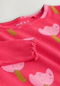 Rózsaszín bordázott hosszú ujjú felső, nagy fehér virágokkal és zöld szárakkal díszítve. Az ujjak vége fodros kialakítású. Márkajelzés látható a nyakkivágás belsejében.