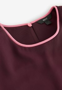 Burgundy top with a smooth texture, featuring a pink trim around the neckline and a pink button closure at the back. Brand tag: NEXT.