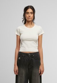 Kremfarget tettsittende t-skjorte med rund hals og korte ermer, prydet med en subtil brodert logo på brystet. Kombinert med mørke, vide jeans.