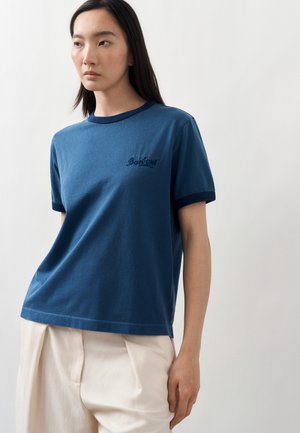 Junge Frau trägt ein blaues T-Shirt mit dunklem Saum und besticktem Text, kombiniert mit lockeren beigen Hosen, vor weißem Hintergrund stehend.