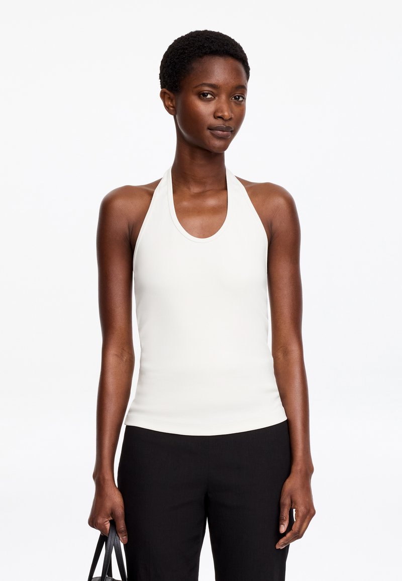 Femme aux cheveux courts et bouclés portant un haut dos nu blanc et un pantalon noir, tenant un sac à main noir avec une expression neutre.