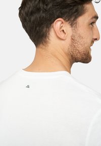 Hvid bomulds t-shirt med rund halsudskæring, der har et lille grå logo på ryggen. Blød tekstur, enkelt design og korte ærmer.