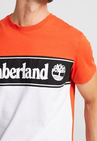 Muž v tričku s krátkým rukávem ve zbarvené kombinaci oranžové a bílé, s černým panelem, na kterém je bílé logo Timberland a emblém stromu.