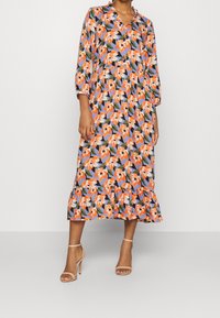Robe midi florale avec un motif orange, bleu et vert vif ; elle possède un col, des manches trois-quarts et un ourlet en couches avec volants.