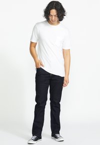 Camiseta blanca de manga corta con un bolsillo en el pecho izquierdo, combinada con jeans de mezclilla oscura y zapatillas bajas negras. Textura suave, ajuste casual.