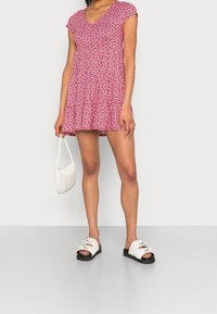 Una mujer lleva un vestido corto de flores rojas, sandalias blancas y sostiene un pequeño bolso blanco frente a un fondo sencillo.