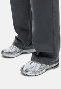 Zilveren sportschoenen met een bovenwerk van mesh en een rubberen zool, gecombineerd met loszittende, donkergrijze broeken. De schoenen hebben witte veters en een gestructureerd ontwerp.