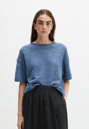 Vrouw met lang bruin haar die een losse blauwe gebreide top met korte mouwen draagt, ingestopt in een zwarte rok met hoge taille, staand tegen een effen achtergrond.