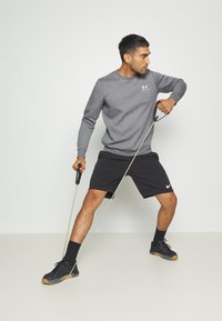 Graues Langarmshirt, schwarze Shorts, mit einem Widerstandsband. Schwarze Sportschuhe mit Gummisohle, stehend vor einem neutralen Hintergrund.