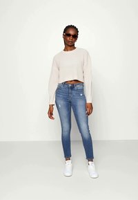Maglione corto beige chiaro, jeans skinny blu con dettagli strappati e sneakers bianche. Gli occhiali da sole completano l'outfit. Trame lisce.