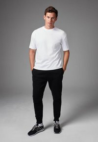 Camiseta blanca de algodón, pantalones jogger negros y zapatillas de running negras con detalles grises. El modelo está de pie con las manos en los bolsillos contra un fondo gris.