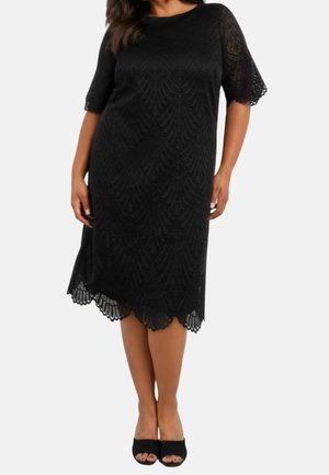 Mujer con un vestido negro de encaje hasta la rodilla, con bordes festoneados y mangas tres cuartos, combinado con tacones abiertos negros.