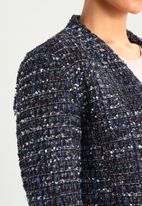 Modrý a černý tweedový blazer s texturovaným tkaním, zdobený růžovými a bílými akcenty a se strukturovaným límcem. Pohled z boku.