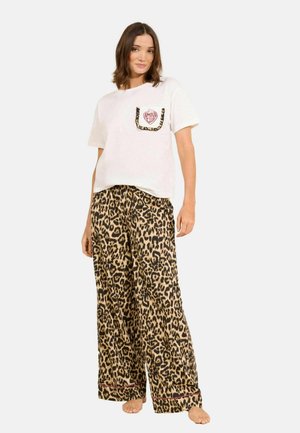 Naine, kes kannab valget T-särki, millel on leopardimustri ääristusega tasku, ja sobivaid laiade säärega leopardimustriga pükse, seisab paljajalu valgel taustal.