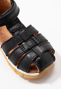 Černá kožená sandál s tkaným designem popruhu, otevřenou špičkou a hnědou vnitřní částí. Disponuje průhlednou bílou podrážkou s texturovanou hnědou základnou.