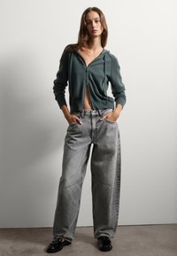 Sudadera verde con cremallera y cordones, llevada sobre unos jeans grises de tiro alto y oversize. El modelo posa con las manos en los bolsillos, de pie frente a un fondo neutro.