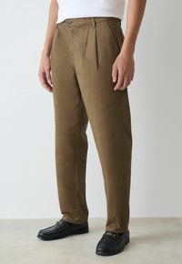 Brown pleated trousers with a fitted waist, made from smooth fabric. Features front pockets and a single button closure. Paired with black loafers.