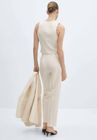 Costum beige din in format din două piese, compus dintr-un top fără mâneci, scurt și o pereche de pantaloni drepți, completat cu o jachetă asortată purtată pe braț.