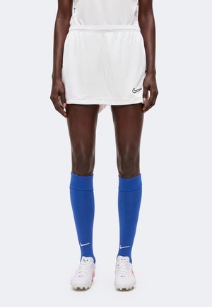 Persona que lleva pantalones cortos deportivos blancos, calcetines altos azules brillantes y botas blancas con detalles negros y naranjas, de pie sobre un fondo blanco.