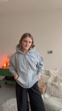 Világosszürke cipzáras kapucnis pulóver puha anyagból, nagy kapucnival, laza fazonnal és oldalzsebekkel. Fekete, széles szárú nadrággal kiegészítve.