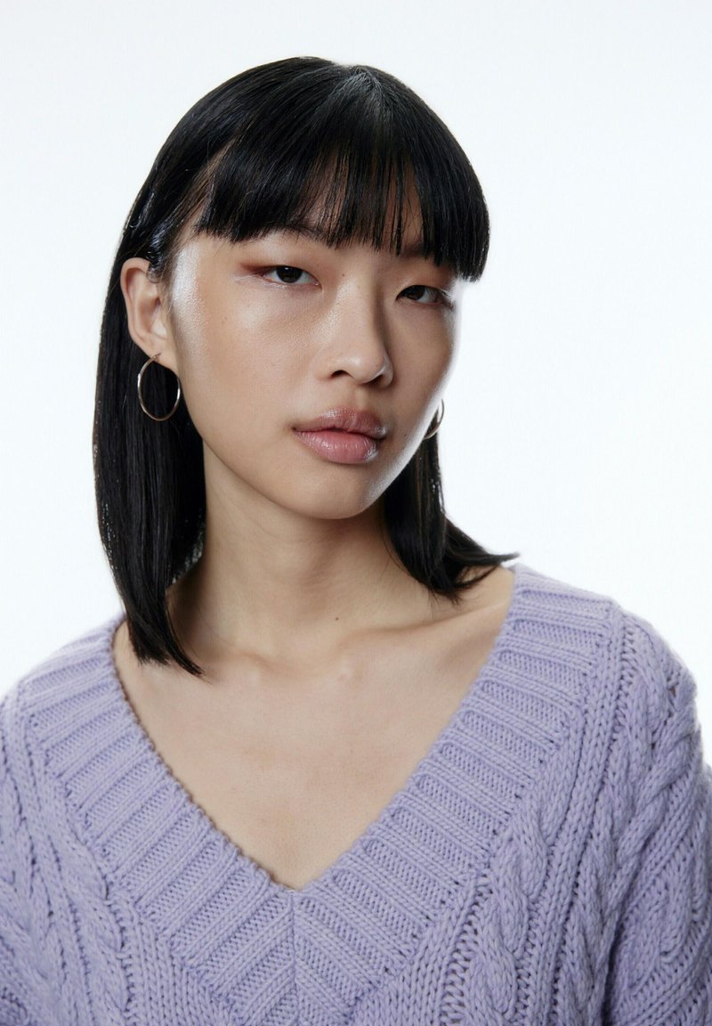 Lavendelfarbener Strickpullover mit V-Ausschnitt und Zopfmuster. Das Model hat glattes schwarzes Haar und trägt silberne Creolen vor einem weißen Hintergrund.