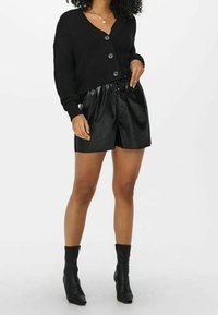 Cardigan noir côtelé avec col en V, fermeture à boutons et manches longues, associé à un short en simili cuir noir et des bottines montantes à la cheville.