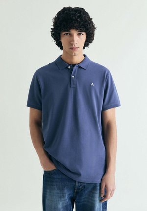 Joven con cabello rizado que lleva una camisa polo azul oscuro y jeans azules, de pie con una mano en el bolsillo frente a un fondo claro.