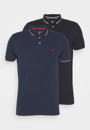 Duas camisolas polo: uma azul-marinho com acabamento vermelho e branco, com logótipo bordado; uma preta com colarinho contrastante. Ambas apresentam uma pala de três botões.