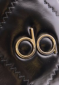 Un material negro brillante presenta un destacado logo dorado "da" con letras estilizadas. La costura visible añade textura a la superficie.