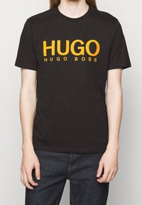 Camiseta de algodón negra con texto naranja en negrita que dice "HUGO" y "HUGO BOSS". Con mangas cortas y cuello redondo. Usada con jeans oscuros.