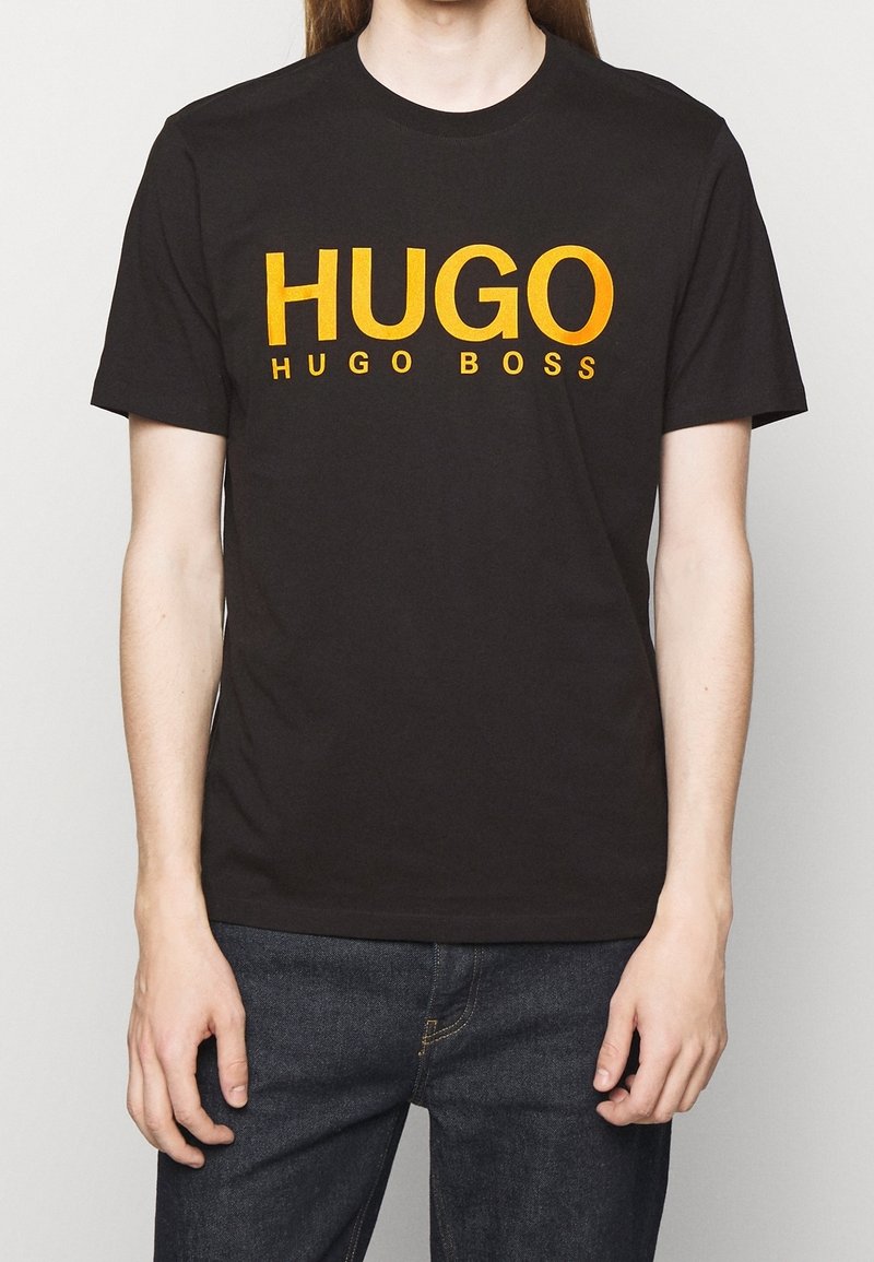 Camiseta de algodón negra con texto naranja en negrita que dice "HUGO" y "HUGO BOSS". Con mangas cortas y cuello redondo. Usada con jeans oscuros.
