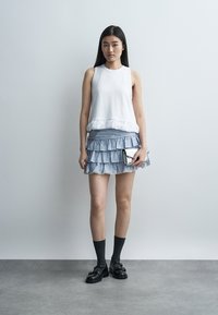 Νεαρή γυναίκα με μακριά σκουρόχρωμα μαλλιά φοράει λευκό αμάνικο τοπ, μπλε μίνι φούστα με βολάν, μαύρες κάλτσες, μαύρα loafers, κρατά ασημένιο τσαντάκι, στέκεται.