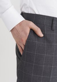 Szare w kratę spodnie z białymi wzorami w siatkę, o teksturowanej tkaninie, z ręką w kieszeni i widocznym rękawem białej koszuli powyżej.