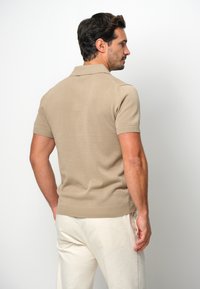 Beige stickad polo tröja med korta ärmar och en revärkrage, bärs med ljusa byxor. Texturerat mönster synligt på tyget.