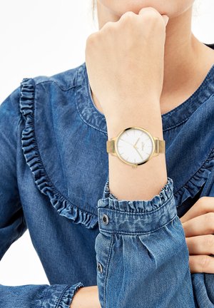 Goldfarbene Armbanduhr mit weißem Zifferblatt, minimalistischen Stundenmarkierungen und Mesh-Armband, getragen an einer Hand mit Jeanshemd-Detail.