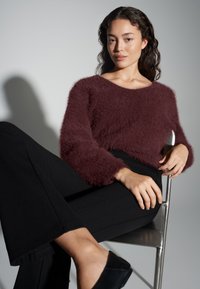 Pull bordeaux duveteux à large encolure et manches longues, associé à un pantalon noir évasé et des chaussures noires, assis sur une chaise argentée.