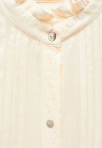 Krémová blůza s perlami na knoflících, špičatým límcem a vertikálním krajkovým panelem podél předního středu.