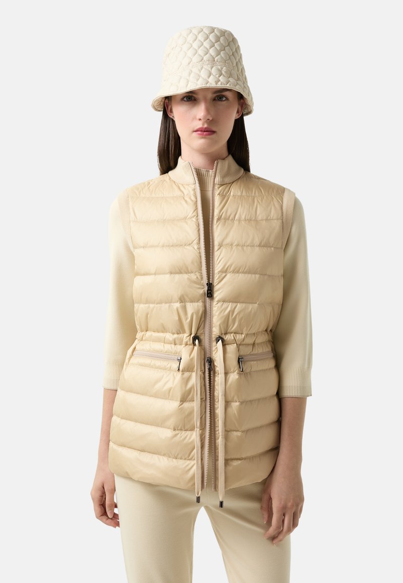 Beige-Steppweste mit hohem Kragen, tailliertem Schnitt und zwei Reißverschlusstaschen. Kombiniert mit einem passenden gesteppte Bucket Hat.