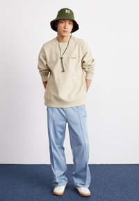 Beige sweatshirt met een klein geborduurd logo, gecombineerd met lichtblauwe trainingsbroek met zijstrepen. Groene bucket hat en witte sneakers.