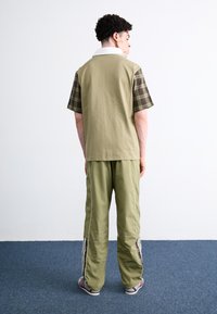 Kurzärmliges grünes und karierter Hemd mit einem weißen Kragen, kombiniert mit grünen Hosen mit seitlichen Streifen. Modell von hinten gesehen.