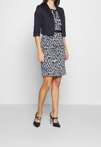 Marineblauwe cropped blazer gecombineerd met een zwart-witte jurk met een patroon van druppelvormen, aangevuld met zwarte hoge hakken.