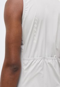 Κοντινό ενός σκουρόχρωμου μπράτσου δίπλα στην πλάτη ενός αμάνικου γκρι ανοιχτού τοπ με την ένδειξη «Isadore» κάθετα τυπωμένη.