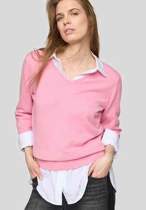 Young woman wearing a pink V-neck sweater over a white button-up shirt with rolled sleeves and dark jeans, hands in pockets.