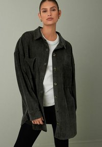 Donkergrijze corduroy shirtjack met een kraag, knopen aan de voorkant en zijzakken, gecombineerd met een witte top en zwarte leggings.