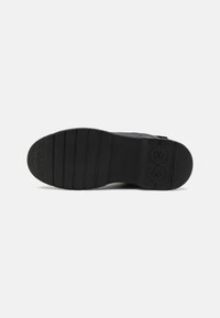 Suela de zapato de goma negra con ranuras horizontales, que presenta un detalle de logo circular y una superficie texturizada para mayor agarre.
