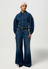 Tuta in denim con maniche lunghe a sbuffo, chiusura con bottoni, due tasche anteriori e gamba larga. Coordinata con una cintura nera aderente e un berretto rosa.