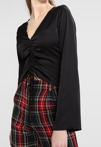 Femme portant un haut noir froncé à manches longues associé à un pantalon taille haute à carreaux rouge et noir, sur un fond uni.