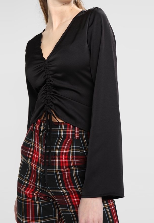 Femme portant un haut noir froncé à manches longues associé à un pantalon taille haute à carreaux rouge et noir, sur un fond uni.